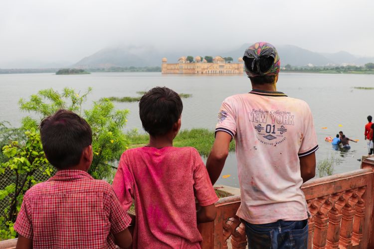 Two boys and one man stand behind a fence and watch men carry a statue into the Jal Mahal lake.