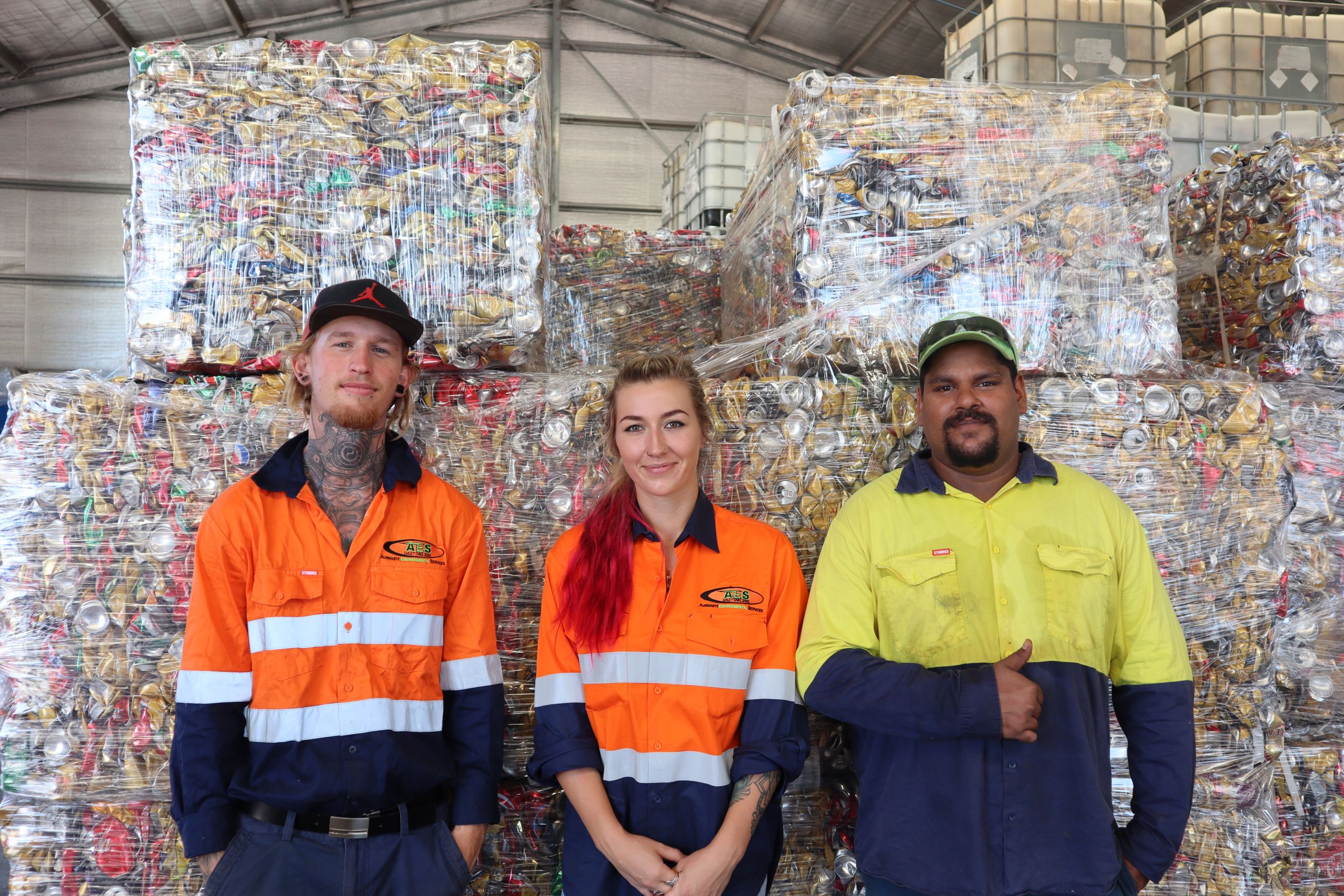 Two men stand side by side a woman. They are in front of wrapped up recycling.